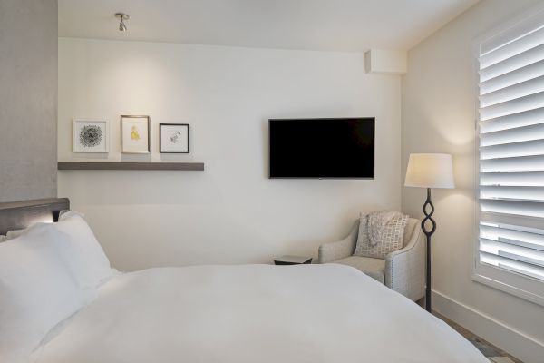 A cozy bedroom with a bed, framed art, a wall-mounted TV, an armchair, a stylish floor lamp, and window shutters.