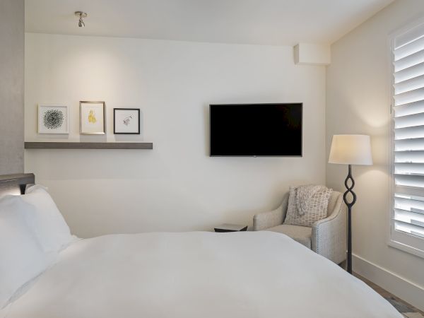 A cozy bedroom with a bed, framed art, a wall-mounted TV, an armchair, a stylish floor lamp, and window shutters.