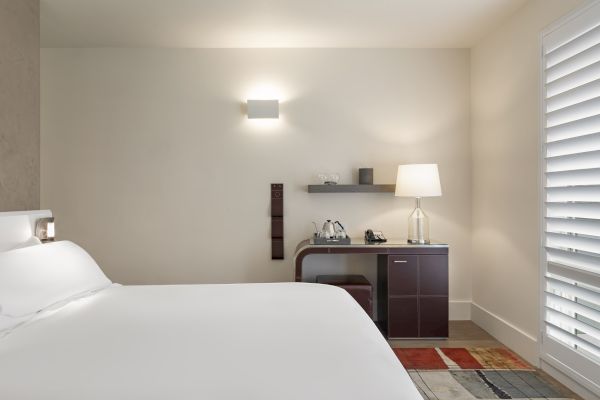 A minimalist hotel room with a white bed, wall light, desk with lamp, kettle, phone, and a window with shutters.