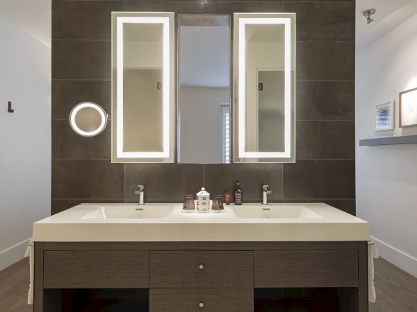 Modern bathroom with dual sinks, illuminated mirrors, and dark tiles. There are toiletries on the counter and framed art on the wall.