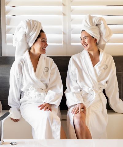 Two people sit on a bathtub edge, wearing white robes and towels on their heads, smiling at each other in a modern bathroom.