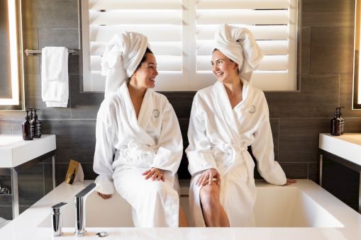 Two people sit on a bathtub edge, wearing white robes and towels on their heads, smiling at each other in a modern bathroom.