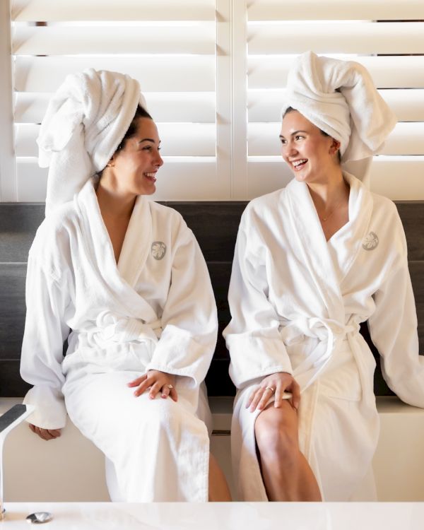 Two people sit on a bathtub edge, wearing white robes and towels on their heads, smiling at each other in a modern bathroom.