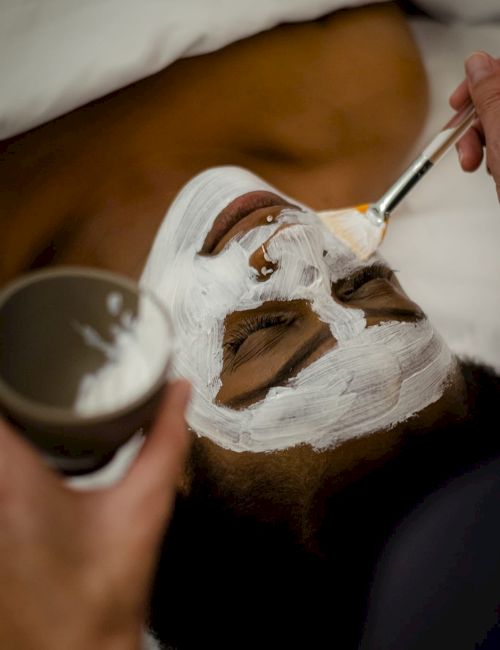 A person getting a facial treatment; white cream is applied with a brush, and they're holding a bowl of the cream.