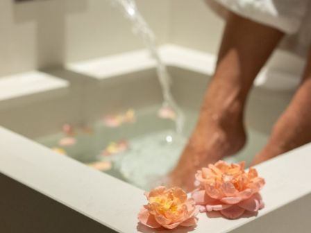 A person in a white robe enjoys a foot bath with flower petals, as water pours from a faucet into the basin.