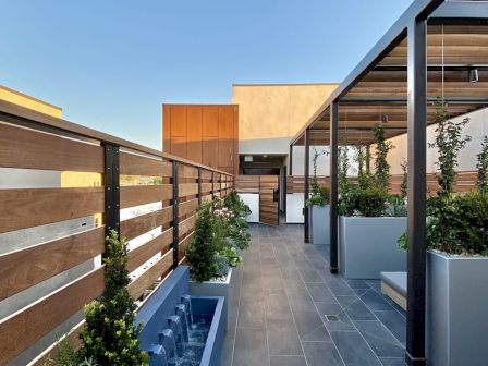 A modern rooftop terrace with a wooden fence, planters, and a pergola under a clear blue sky.