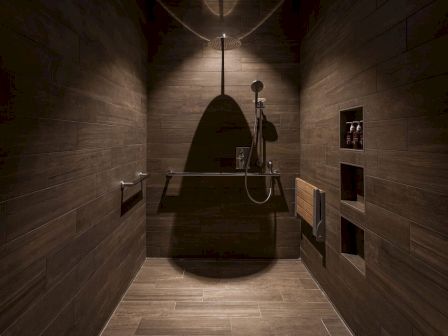 A modern, dimly lit shower with dark tiles, handheld showerhead, bench, and grab bars, creating a sleek and minimalist atmosphere.