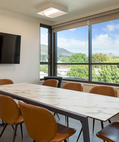 A modern conference room with a marble table, brown chairs, wall-mounted TV, large window with a scenic view of trees and hills outside.