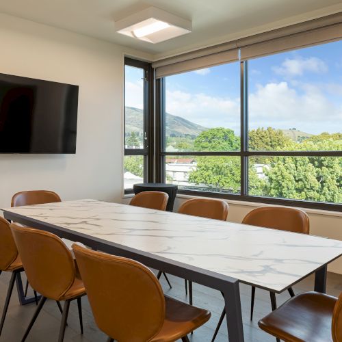 A modern conference room with a marble table, brown chairs, wall-mounted TV, large window with a scenic view of trees and hills outside.