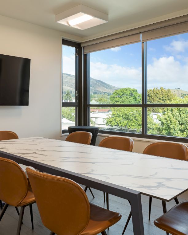A modern conference room with a marble table, brown chairs, wall-mounted TV, large window with a scenic view of trees and hills outside.