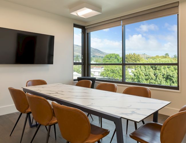 A modern conference room with a marble table, brown chairs, wall-mounted TV, large window with a scenic view of trees and hills outside.