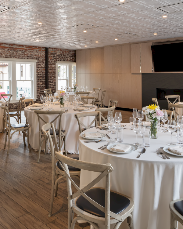 An elegantly set dining room with round tables, white linens, and simple floral centerpieces, suitable for events or gatherings.