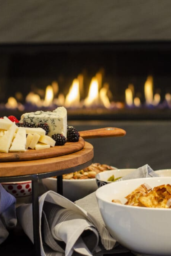 A cheese platter with berries, strawberries, and bread sits near a cozy fireplace, creating a warm and inviting atmosphere.