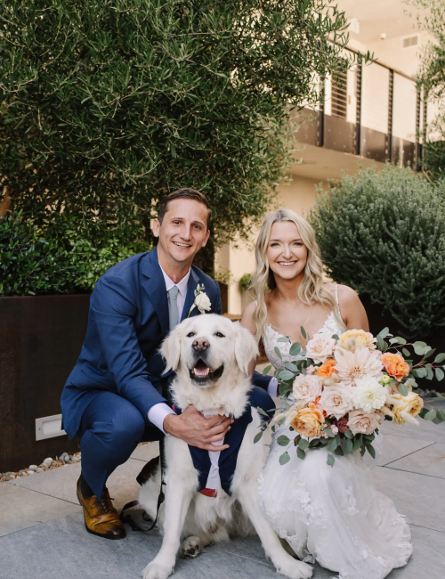 A couple in wedding attire poses with a dog in a suit, holding a bouquet, surrounded by greenery.