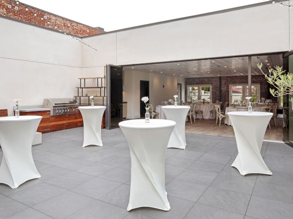 An outdoor event space with cocktail tables, covered with white cloths, adjacent to an indoor area with dining tables and chairs.