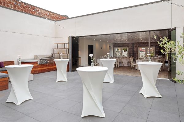 The image shows an outdoor event space with tall tables, white tablecloths, and simple flower centerpieces.