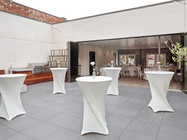 The image shows an outdoor event space with tall tables, white tablecloths, and simple flower centerpieces.