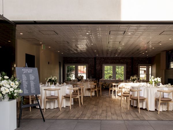 A decorated venue with round tables, white tablecloths, chairs, flowers, and a menu board, set for an event in a spacious room.