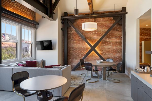 A modern living area with exposed brick, steel beams, cozy seating, a round dining table, and a wall-mounted TV by large windows.