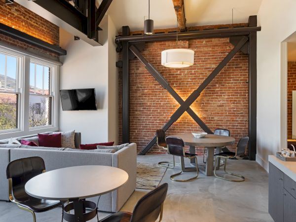 A modern living area with exposed brick, steel beams, cozy seating, a round dining table, and a wall-mounted TV by large windows.