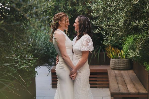 Two people in wedding dresses are holding hands and smiling at each other, surrounded by lush greenery outdoors.