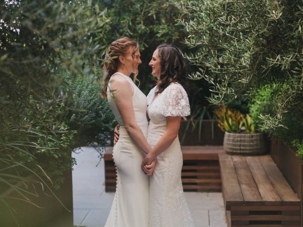 Two people in wedding dresses are holding hands and smiling at each other, surrounded by lush greenery outdoors.
