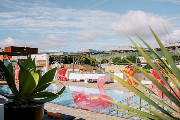 A rooftop pool scene with pink flamingo floats, green plants, tables with red covers, and distant hills under a partly cloudy sky.