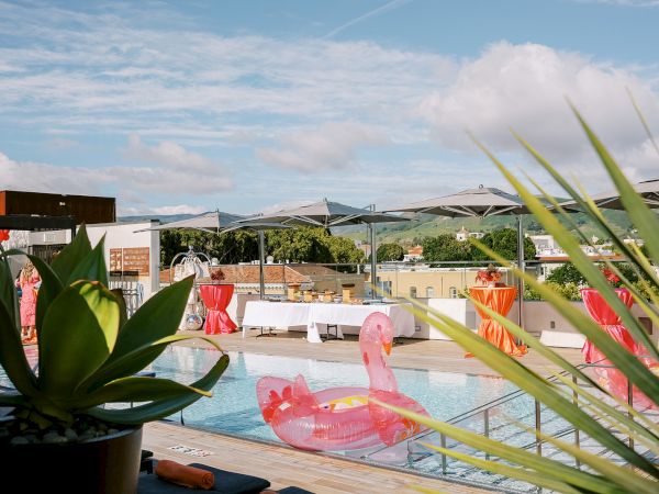 A rooftop pool scene with pink flamingo floats, green plants, tables with red covers, and distant hills under a partly cloudy sky.