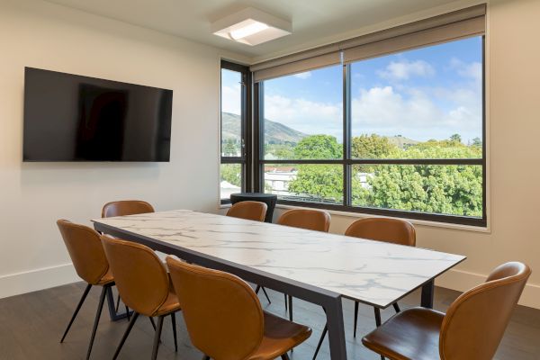 A modern conference room features a marble table, brown chairs, a wall-mounted TV, and a large window with a scenic view.