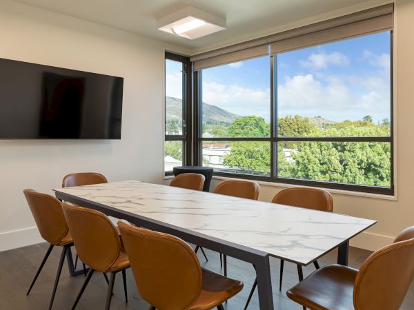 A modern conference room features a marble table, brown chairs, a wall-mounted TV, and a large window with a scenic view.