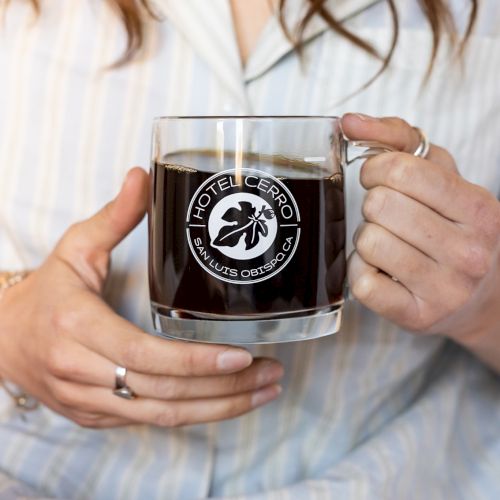 A person holding a clear mug with black coffee labeled "Hotel Cerro San Luis Obispo CA," wearing a striped shirt and rings.