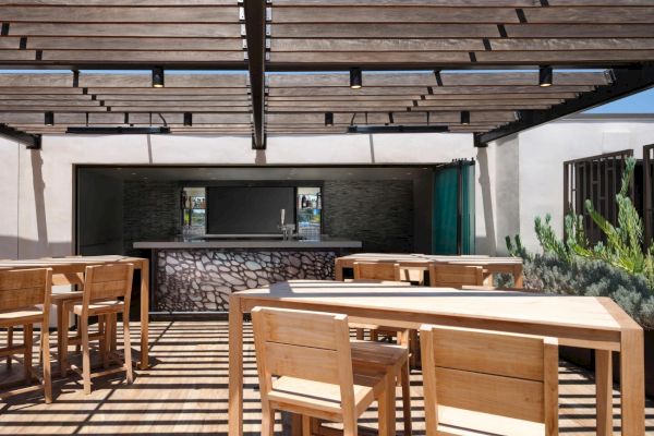 An outdoor dining area with wooden tables and chairs under a pergola, next to a bar with a stone design.