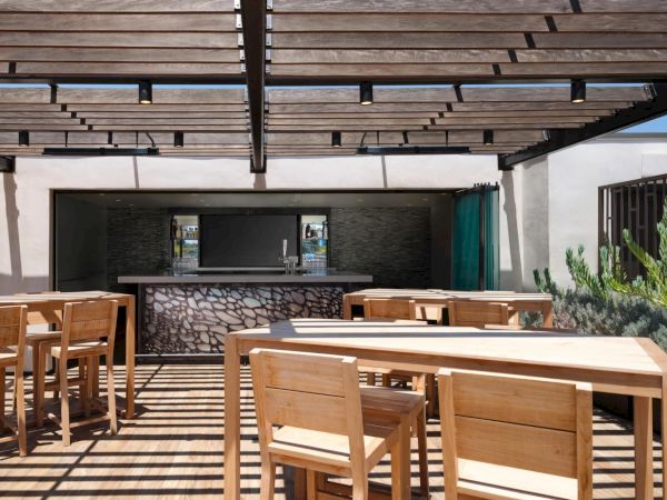 An outdoor dining area with wooden tables and chairs under a pergola, next to a bar with a stone design.