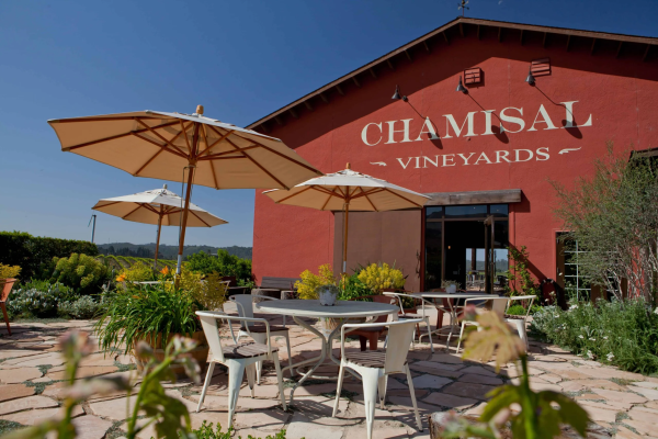 Outdoor seating with umbrellas at Chamisal Vineyards, featuring a red building and surrounding greenery under a clear blue sky.