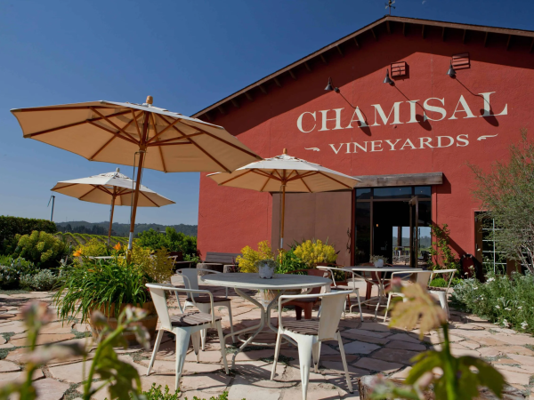 Outdoor seating with umbrellas at Chamisal Vineyards, featuring a red building and surrounding greenery under a clear blue sky.