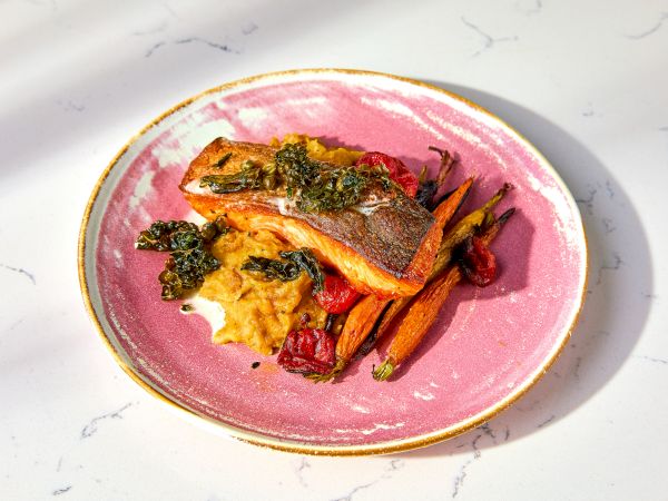 Seared salmon on a pink plate with mashed vegetables, roasted carrots, and a garnish of greens.