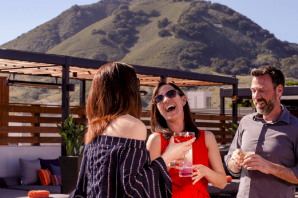 Three people outdoors are laughing and chatting, each holding a drink, with a mountain visible in the background.