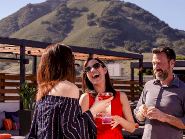Three people outdoors are laughing and chatting, each holding a drink, with a mountain visible in the background.