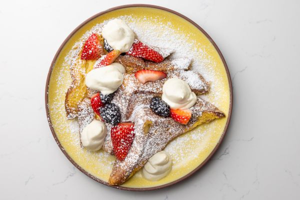 French toast with whipped cream, strawberries, blackberries, and powdered sugar on a yellow plate.