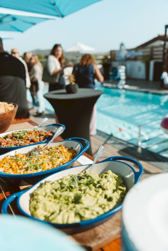 Poolside outdoor party with a buffet of colorful dishes on a table, while guests mingle in the background near the blue water.