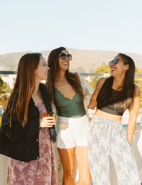 Three women stand on a sunny patio, wearing sunglasses and smiling and laughing together while holding drinks.