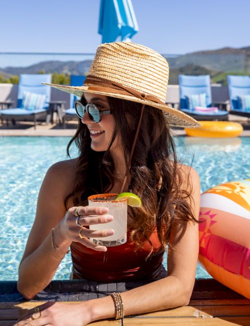 A person wearing a straw hat and sunglasses enjoys a drink by the pool, with a colorful float and loungers in the background.