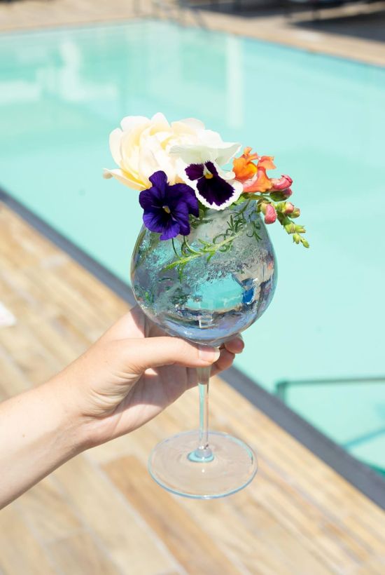 A hand holds a decorative wine glass filled with flowers and foliage against a backdrop of a wooden deck and swimming pool.