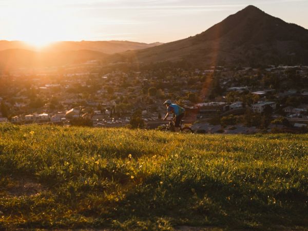 A cyclist rides on a grassy hill at sunset, with mountains and a town in the background.