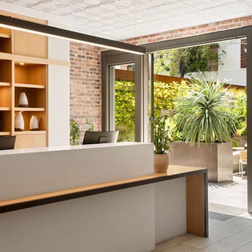 A modern indoor reception area with wooden shelves, plants, and natural light streaming through glass doors to a lush outdoor patio.