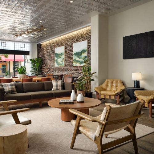 A cozy, modern lounge with wooden furniture, plants, a fireplace, artwork, and large windows letting in natural light.