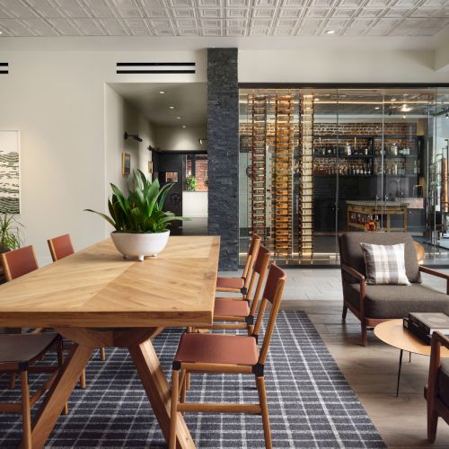 A modern interior with wooden furniture, a large plant, artwork on the wall, and a glass wine display in the background.