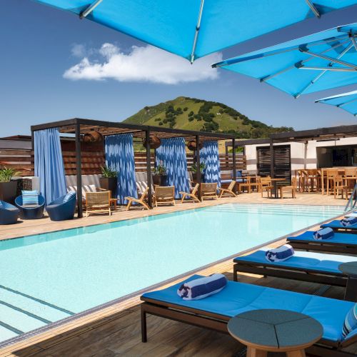 A luxurious pool area with blue loungers, cabanas, and umbrellas, surrounded by wooden deck, chairs, and a scenic hill view under a clear sky.