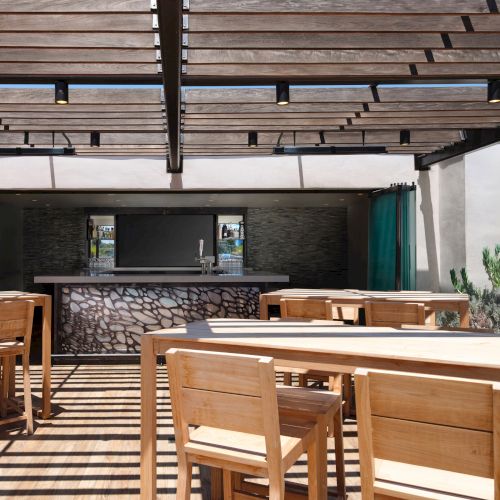 Outdoor seating area with wooden tables and chairs under a pergola, and a bar with a decorative stone front.