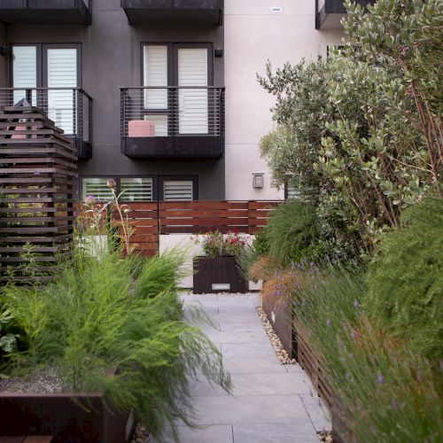 A modern garden with lush greenery, pathway, wooden structure, and a building with balconies in the background.